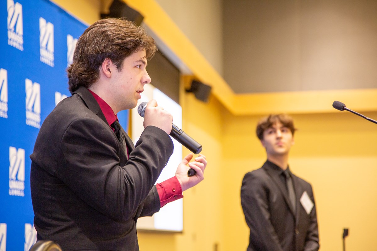 A person holds a microphone and makes a presentation while another person looks on.